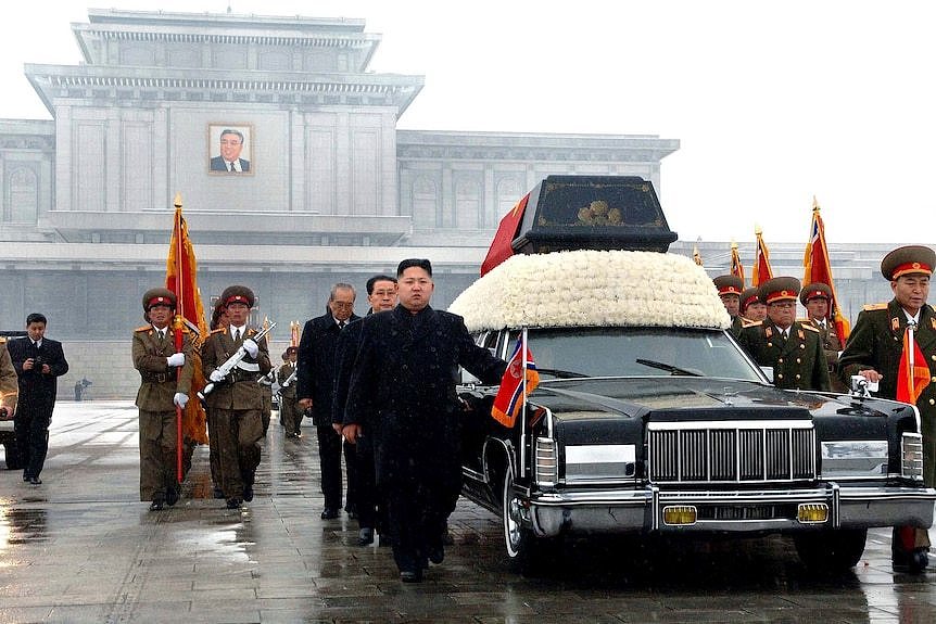 North Korean soldiers and other men walk beside a black car with flowers on it.