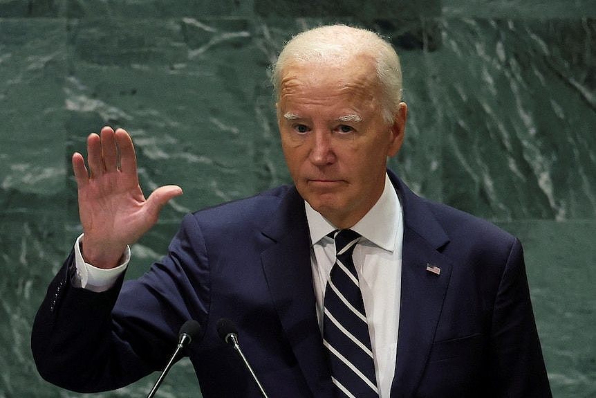 Joe Biden stands in front of a green wall and holds up a hand.