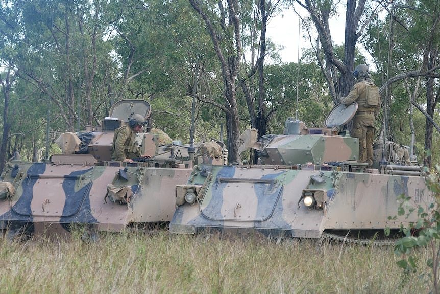 A handful of uniform soldiers stand on top of two military tanks in bushland