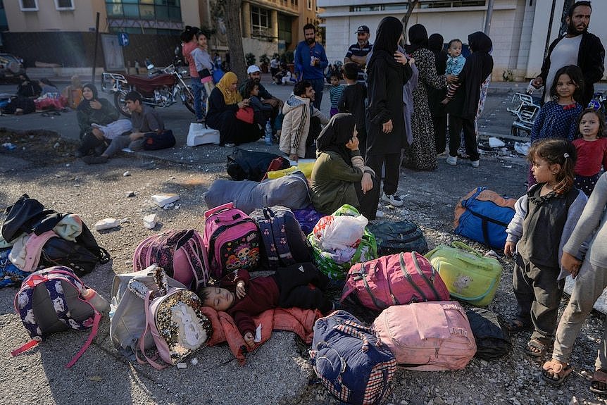 A bundle of bright backpacks sits on the ground with a child lying down in the middle in front of a group of huddled people