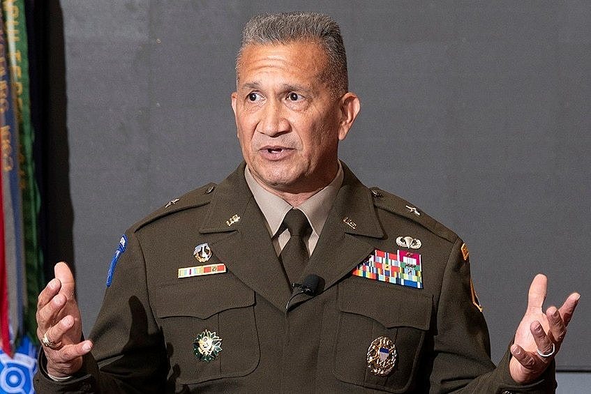 A man with a short tight haircut and military uniform stands speaking.