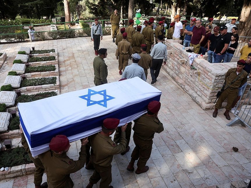 Six soldiers in uniform carry a coffin draped in an Israeli flag toward a group of mourners in a cemetery