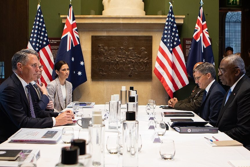 Several people sit around a table, with Australian and US flags behind them.