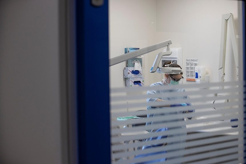 Looking through the door of a dental clinic where a dentist inside prepares a chair and equipment.