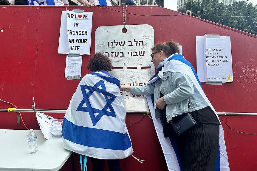 Two people stands at a memorial of names of hostages written on a large dog tag.