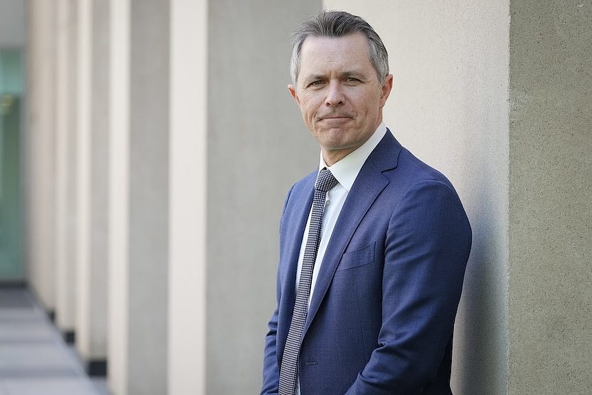 Jason Clare, wearing a suit, stands against a wall. 