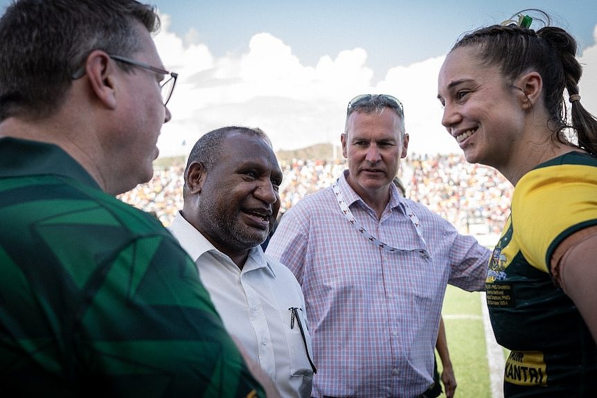 A Polynesian man smiles as he speaks to a NRLW player as two white Australian men look on at a footy field