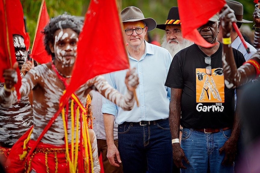 White man with hat walks in the middle of a group of Indigenous people in traditional dress carrying red flags