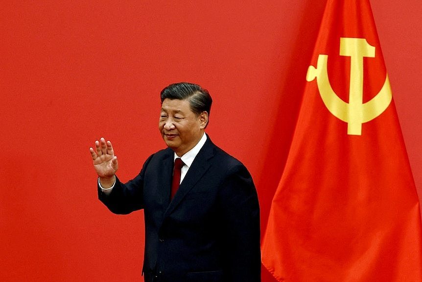 Xi Jinping gives a little wave next to a Chinese flag