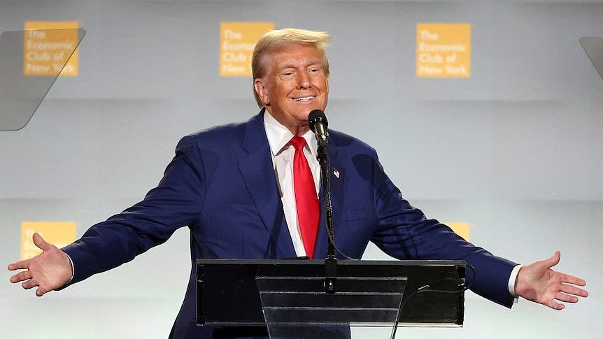 Donald Trump stands at a podium, holds his hands outstretched and smiles.