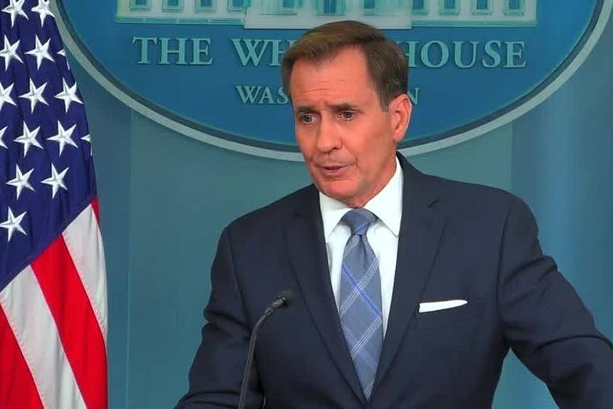 A man in a suit and tie stands in front of a microphone with an American flag next to him