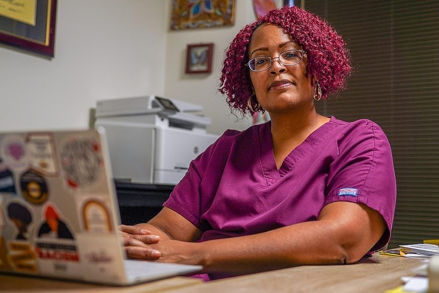 DeShawn Taylor, with dyed purple hair and purple scrubs, sits behind a laptop with colourful stickers on it.