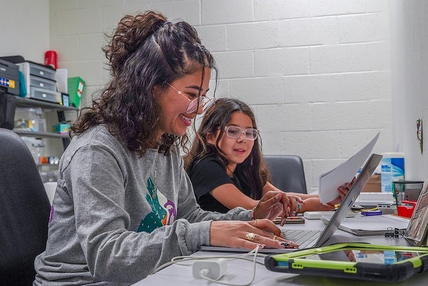 A woman sits with her young daughter at a desk, looking at a laptop computer.