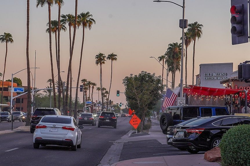 Cars drive down a city street past shopfronts and palm trees at dusk.