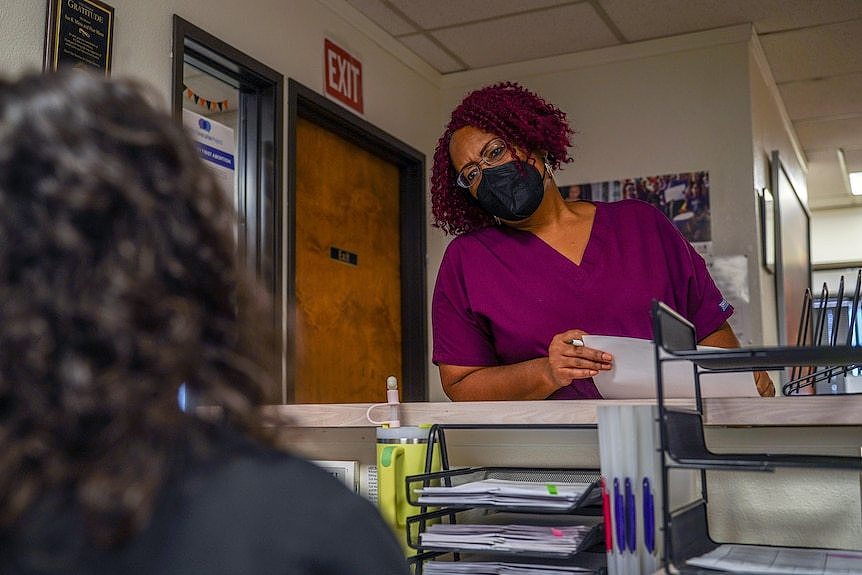 A woman in scrubs and a mask talks to another woman behind a desk