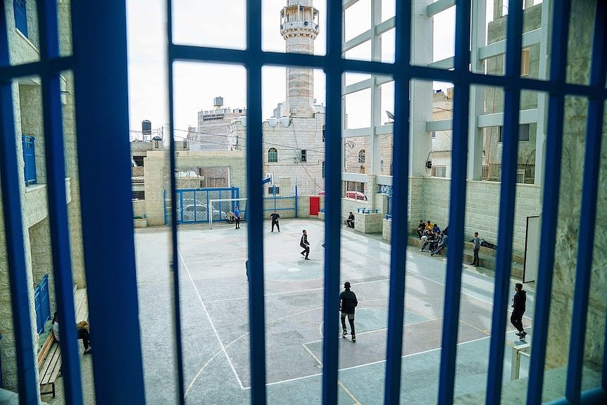 Children play soccer in a concrete field behind bars with a guard tower in the background