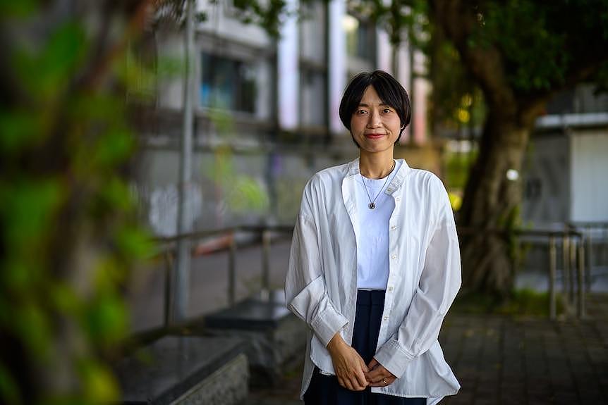 A woman with short hair waring a white shirt stands outside in the shadow of a tree