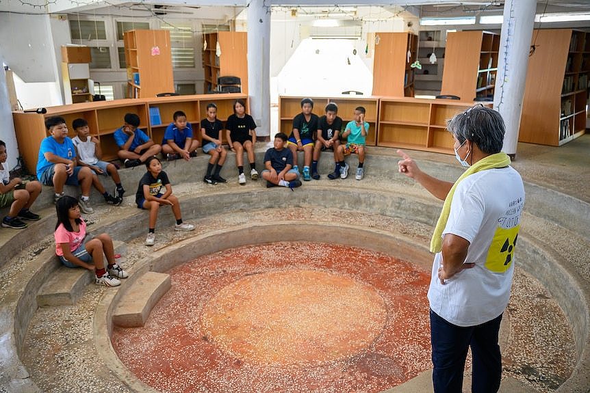 A man stands in the centre of a circular room with younger kids sitting on the steps