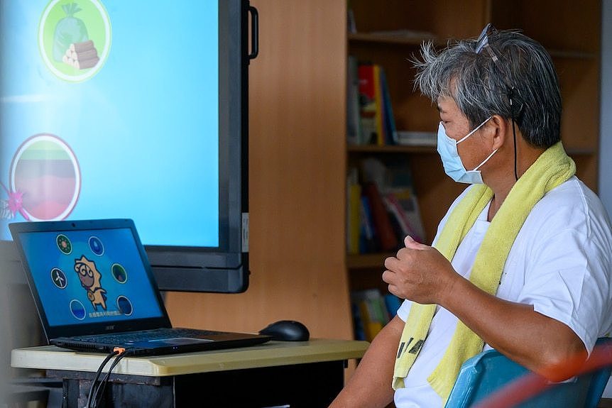 A man wearing a face mask and a scarf sits on a chair. a laptop is open in front of him