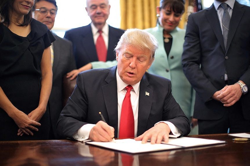 Donald Trump signs a page as a number of people stand around him 