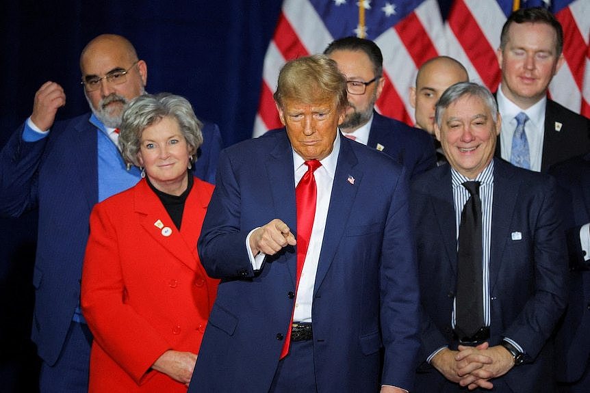 Donald Trump stands on stage with his team, pointing at crowd