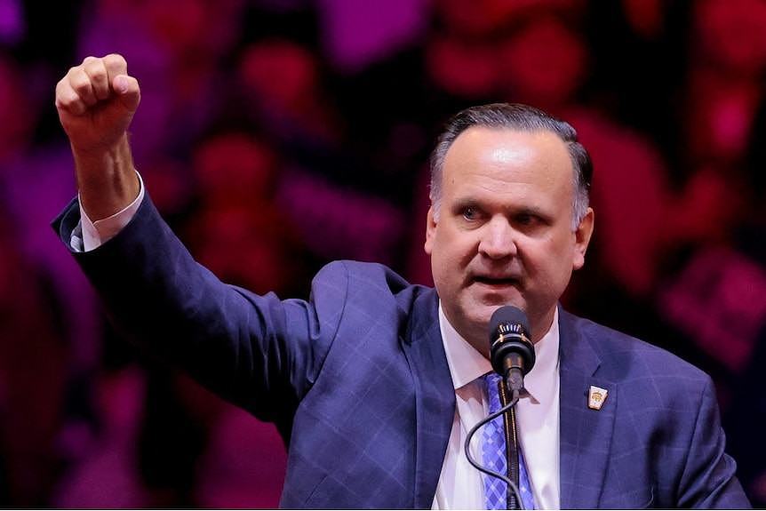 A man in a suit speaks into a microphone while raising his hand in the air