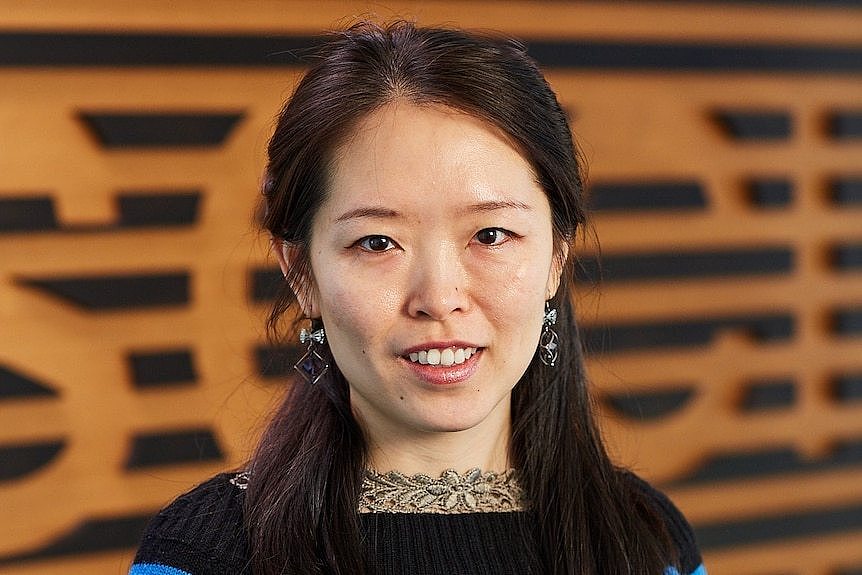 A woman in a blue top stands in front of a wooden wall.