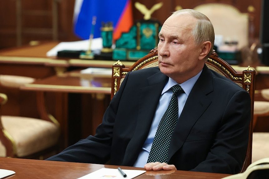 Vladimir Putin wearing a dark suit, tie and blue shirt sitting emotionless at a wooden desk in front of a Russian flag