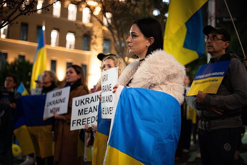 A woman with dark hair standing draped in a blue and yellow Ukrainian flag alongside protesters holding white placards