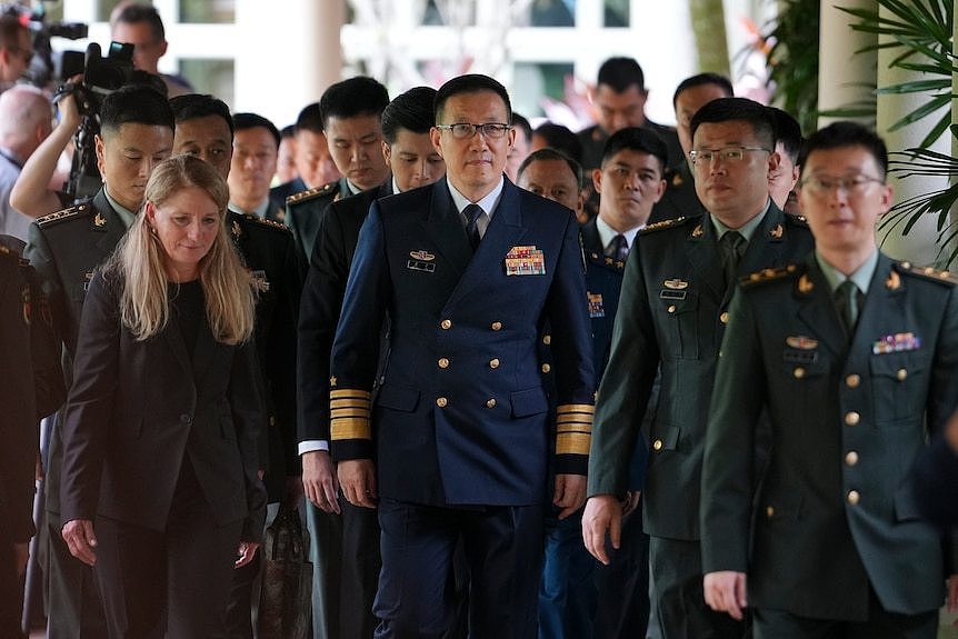 A crowd of people, mostly Chinese men in military uniform, walk down a corridor. The man in the centre is wearing blue.