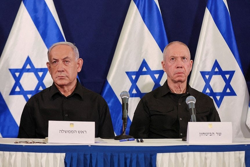 Two men sitting at a table, with flags behind them, looking on