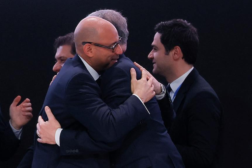 COP29 president Mukhtar Babayev embraces another man while smiling and wearing a dark suit