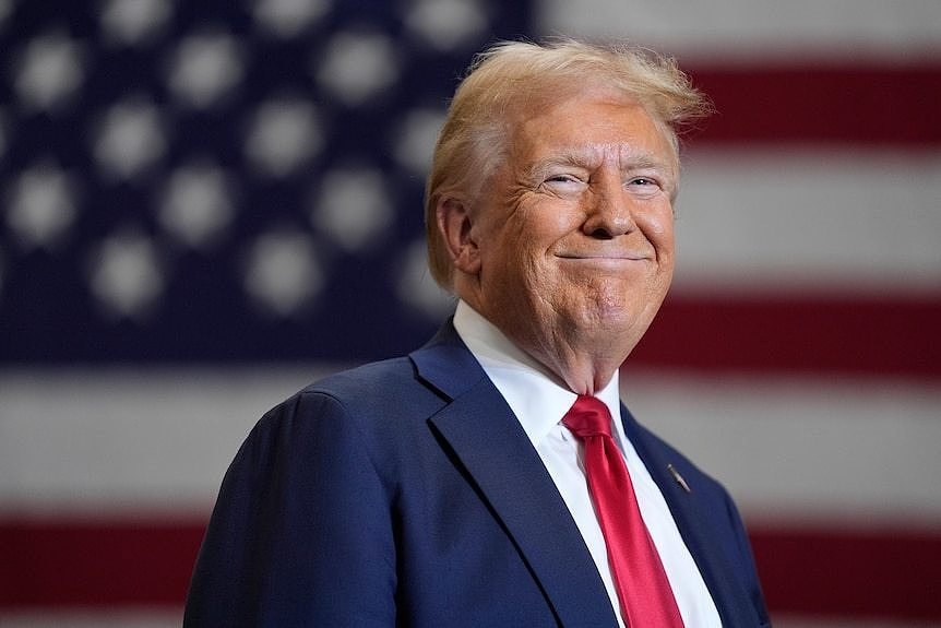 Donald Trump smiles as he stands in front of an American flag.