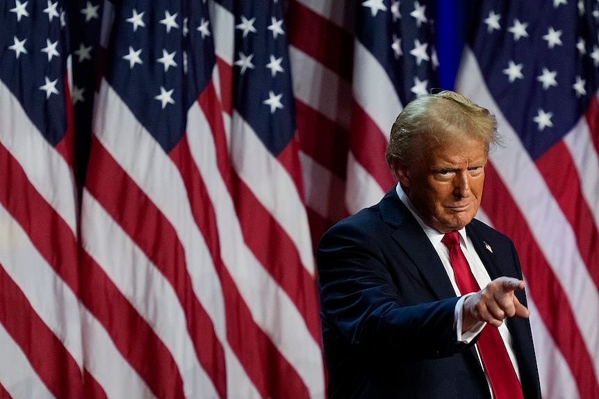 Donald Trump points to the crowd while standing on stage surrounded by United States flags.