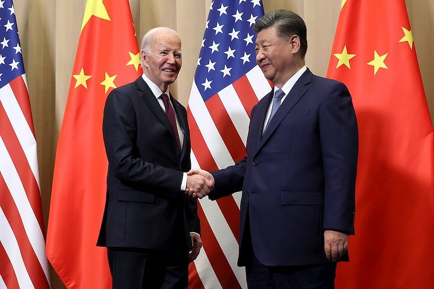 Joe Biden and Xi Jinping wearing dark suits and shaking hands in front of US and Chinese flags