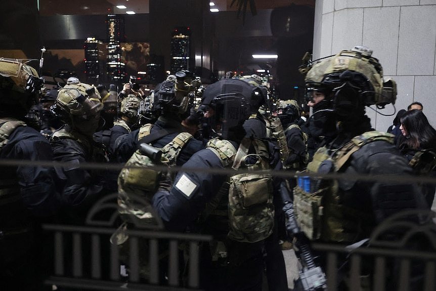 Soldiers stand in a huddle outside a building in camouflage gear and protective wear 