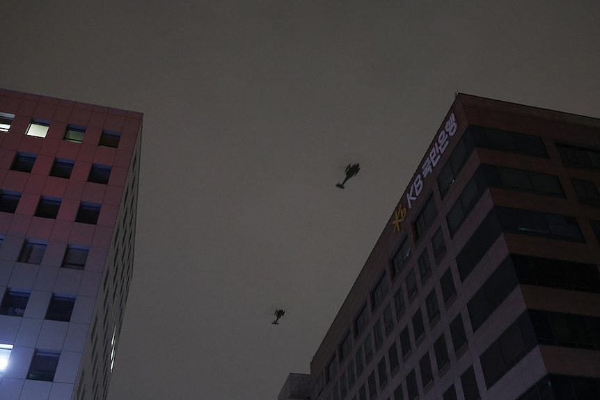 Two helicopters fly above multi-storey buildings against a dark grey sky