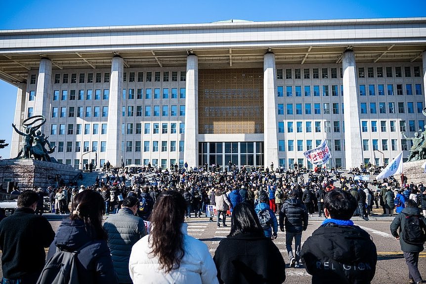 Hundreds of people gather on the steps of a large columned parliamentary building during the day