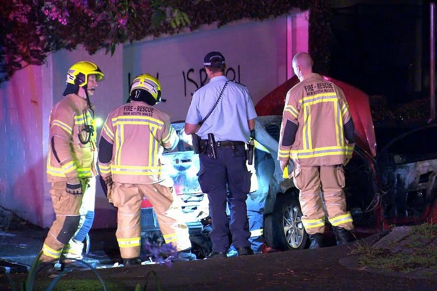 Police and firefighters look at smouldering car in Woolahra after anti-Israel graffiti visible in fence behind