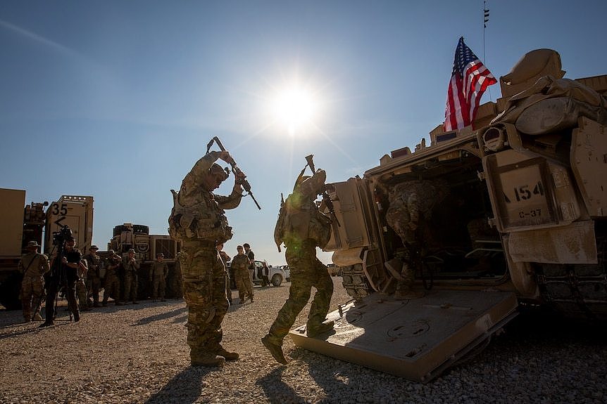 US troops carrying gunswalk towards a combat vehicle flying an American flag.