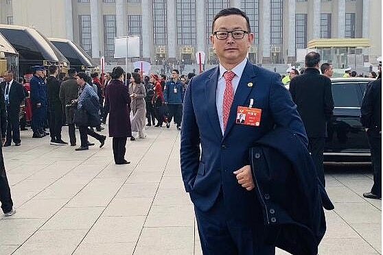 A man in a suit wearing a red badge looks neutrally at the camera in front of a large concrete building