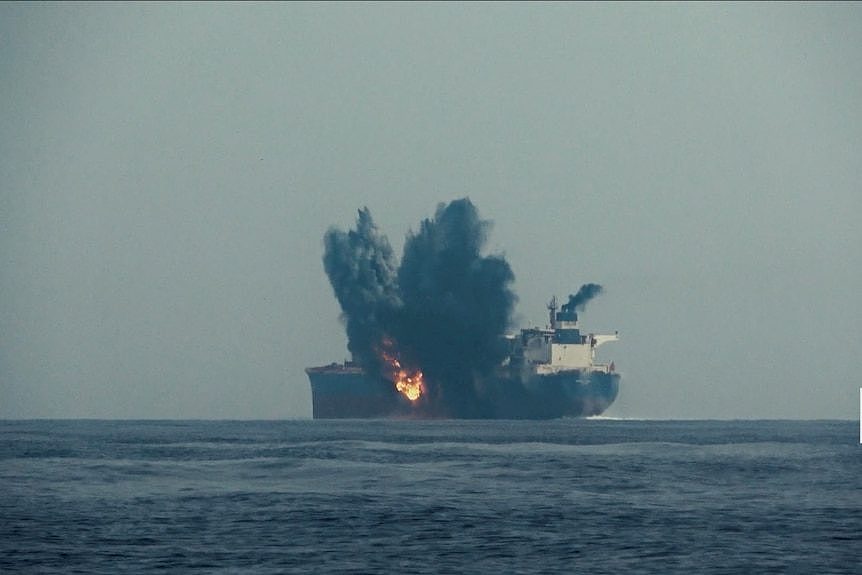 A large tanker ship in open ocean is on fire with black smoke pouring from it