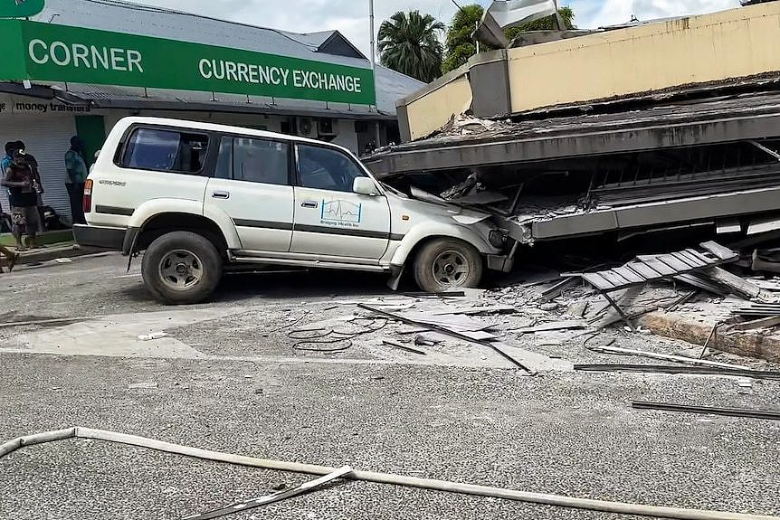 A silver four-wheel-drive vehicle with its front end squashed under a collapsed building roof with debris scattered around