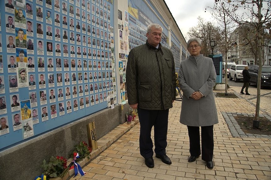 A man in a green long-coat stands next to a woman in a long grey coat alongside a wall with pictures of people on it