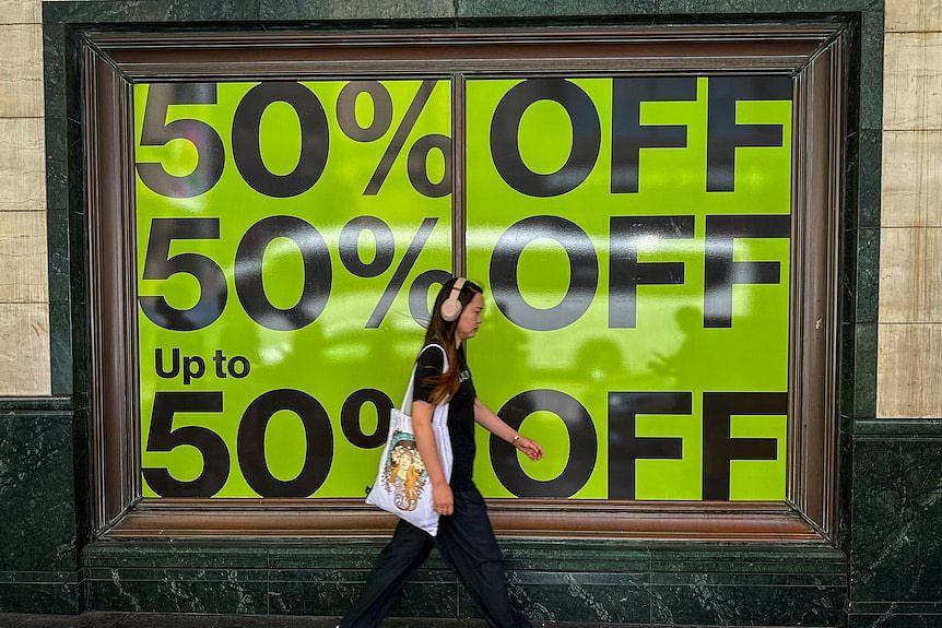 A woman with beige headphones and a tote bag walks past a shop window advertising 50 per cent off.