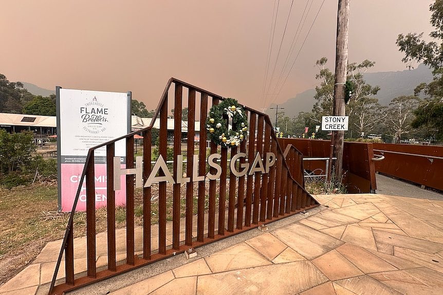 'Halls Gap' sign in silver letters, on rusty, artistic sign in the street, in front of a wall of smoke.