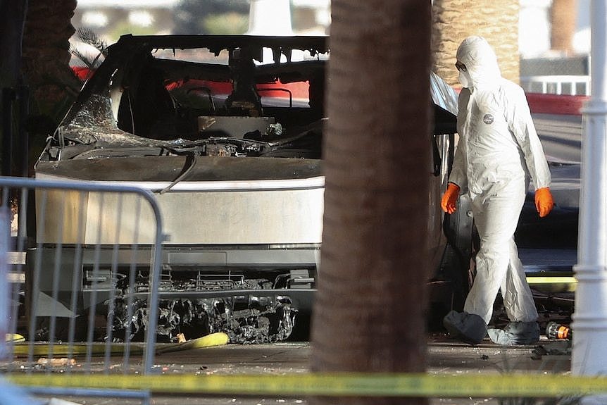 A person wearing a HAZMAT suit inspects the burnt-out body of a Cybertruck