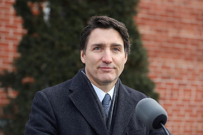 Justin Trudeau, dressed in a black winter coat, stands at a microphone in front of a red-brick wall with greenery on it.