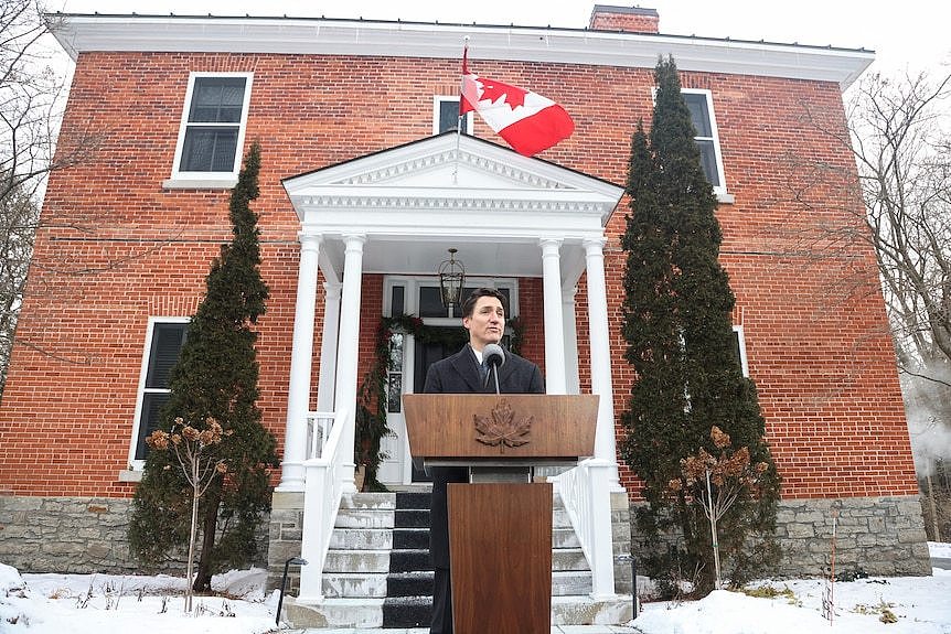 Justin Trudeau, dressed in a black winter coat, stands at a podium in front of a red-brick cottage with a Canadian flag on it.