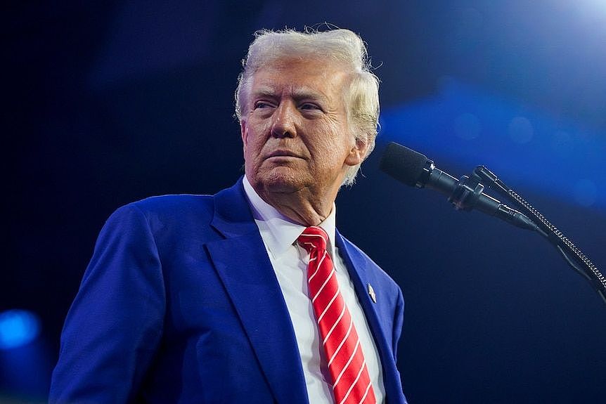 Donald Trump wears a blue suit and red tie as he looks to his side while standing at a podium